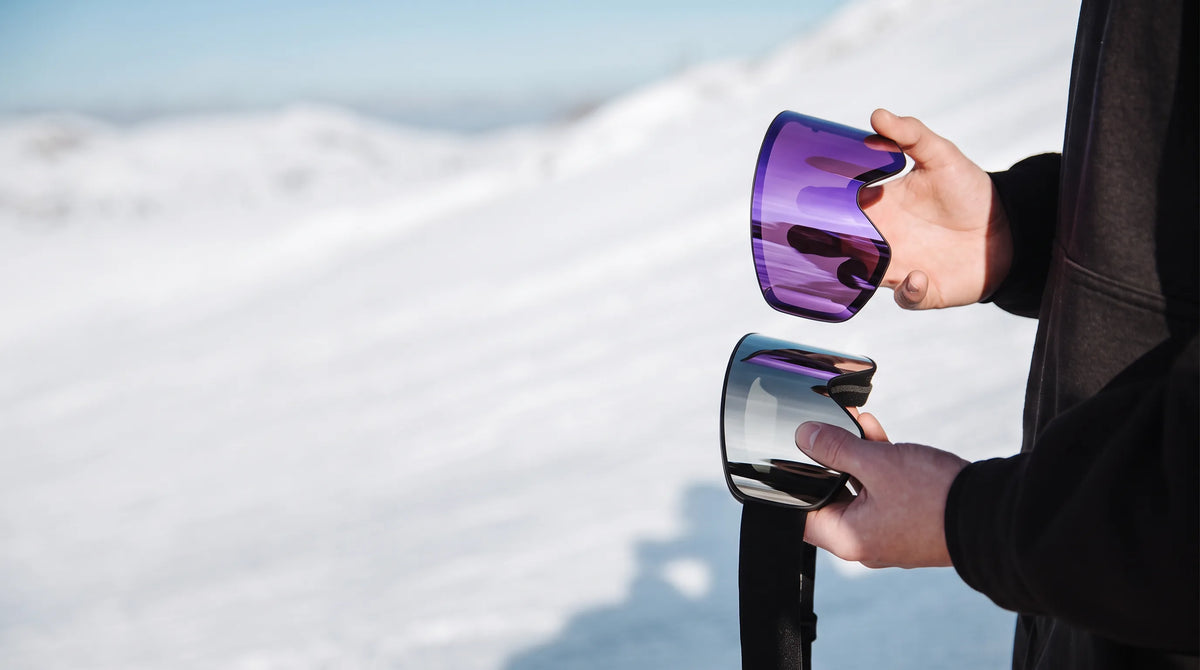 Hands swapping a purple magnetic visor onto a goggle on a snowy slope
